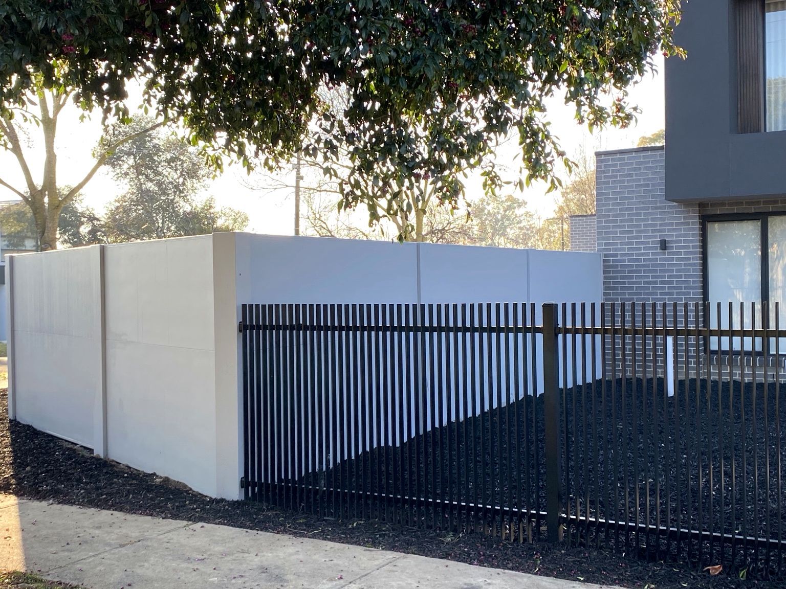 A pale grey VogueWall boundary with black aluminium blade fencing in front of it, under a street tree, Ashburton.