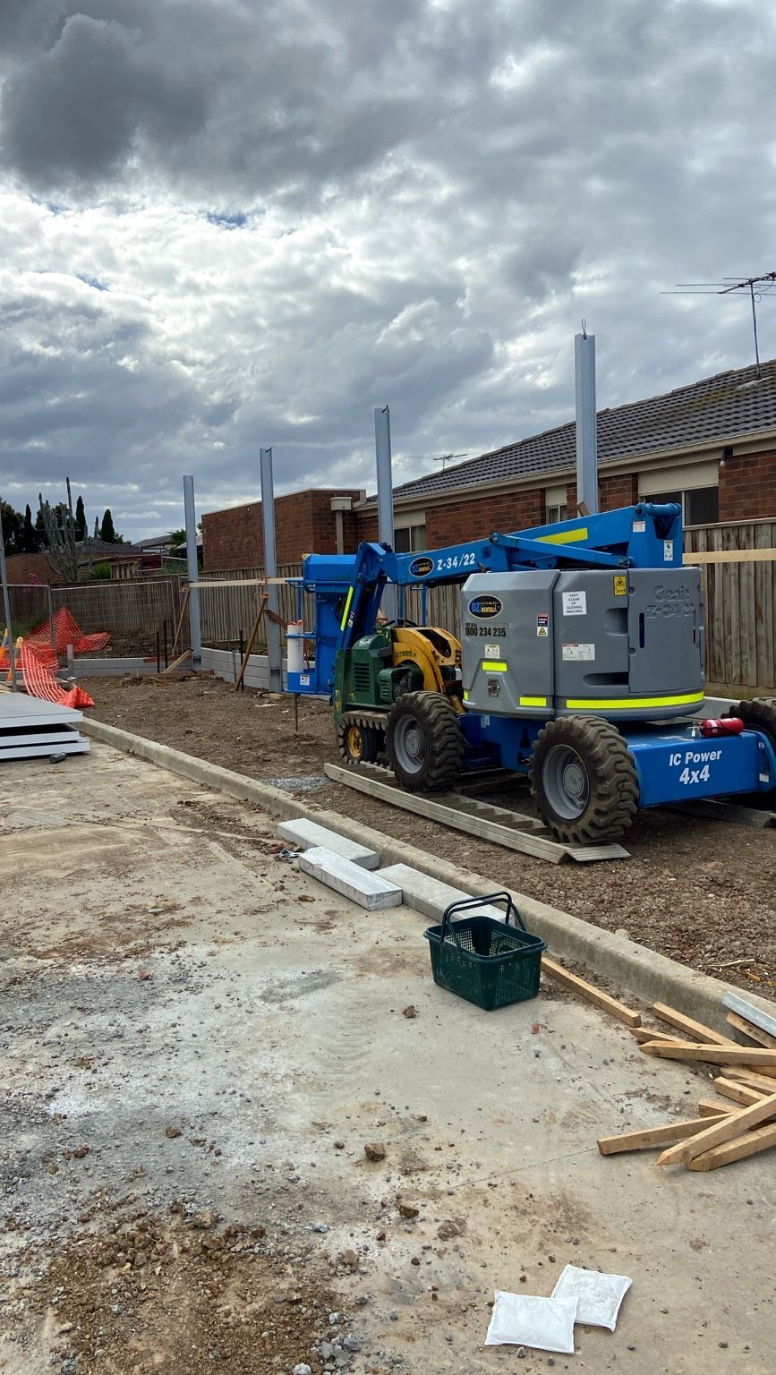 Steel posts standing at Point Cook ahead of the panels, a work platform beside them and houses beyond the timber fence.